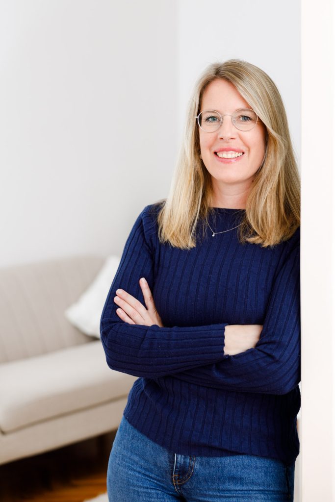 Eine Frau in einem blauen Pullover und mit Brille steht vor einem Sofa und vermittelt den Eindruck einer entspannten Atmosphäre.